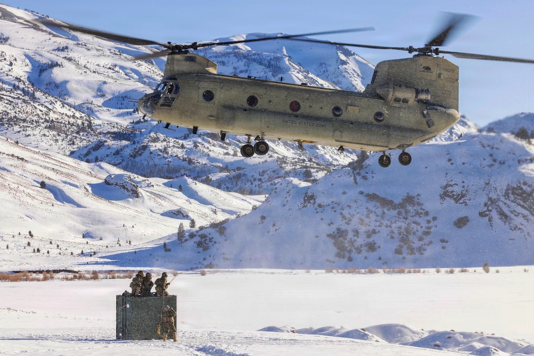 A helicopter with two propellers hovers over three people kneeling on a box and another person standing in the snow, with snow-covered mountains in the background.
