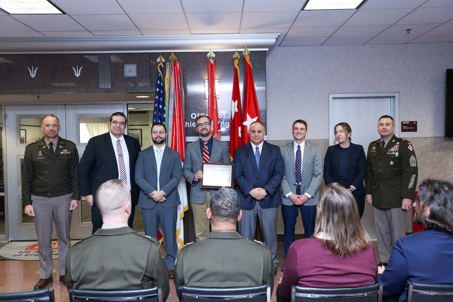 A group of individuals stand shoulder to shoulder in front of individuals who are seated. There are flags in the background.