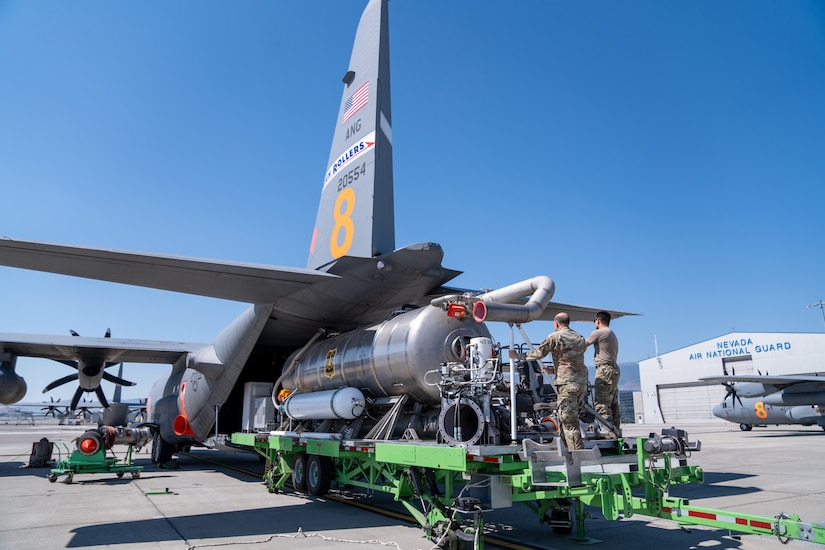 Two men in camouflage military uniforms stand on an equipment loader as they guide a large firefighting system into the back of an open military aircraft that is sitting on a tarmac. An aircraft hangar is in the background, with the words “Nevada Air National Guard” written on the front.