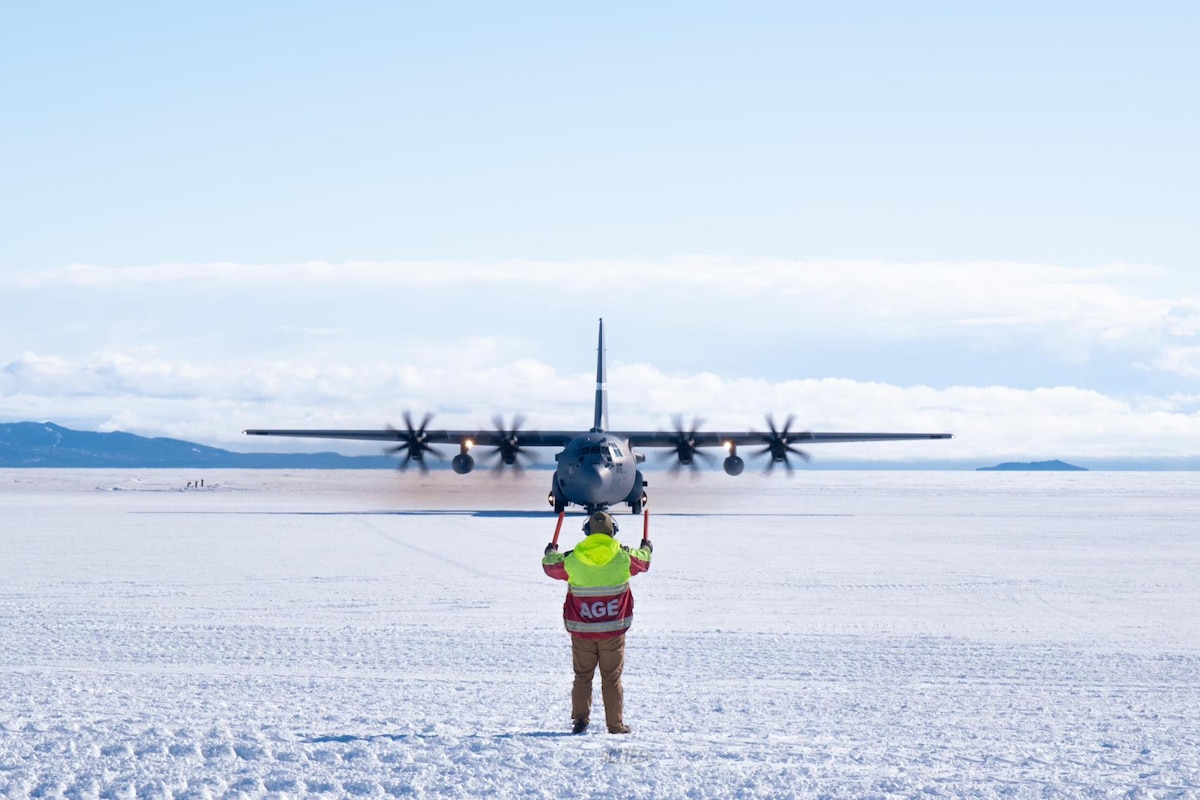 A person in winter attire marshals in a large military aircraft on a snowy plain. There are mountains in the distant background.