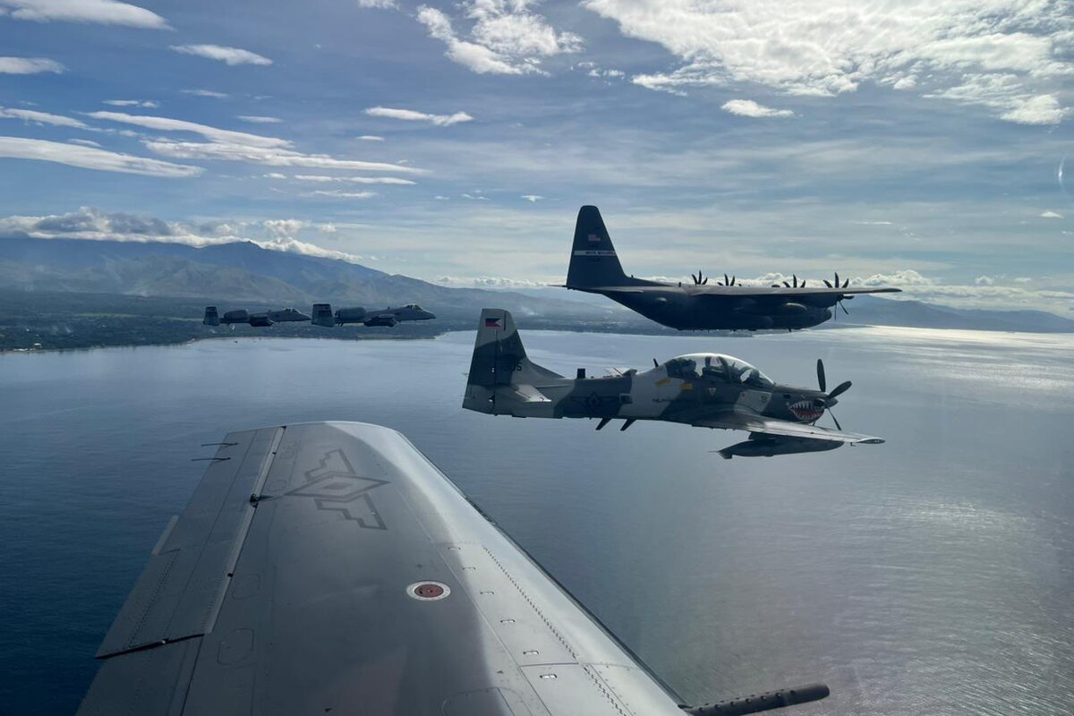 Five military aircraft fly in formation over a large body of water. The coastline is the background with mountains in the distance.