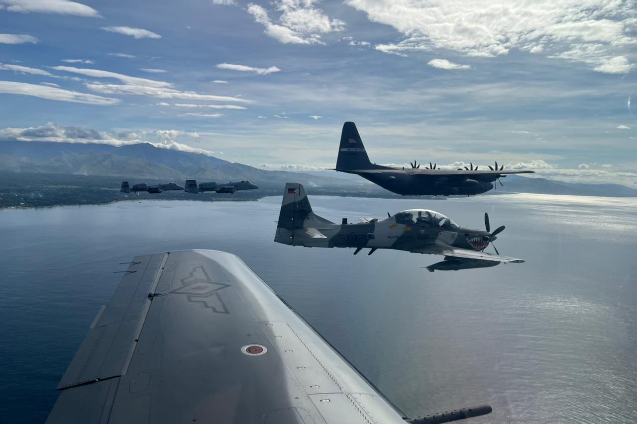 Five military aircraft fly in formation over a large body of water. The coastline is the background with mountains in the distance.