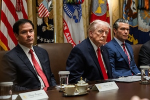 Three men in business suits sit at a long table, with flags in the background.