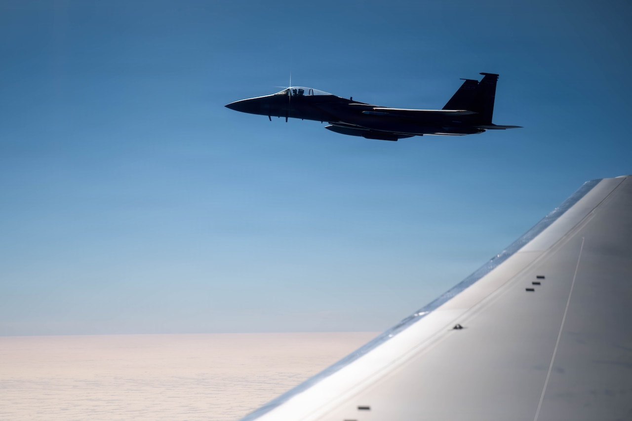 A military jet flies through the air. The wing of another military aircraft is in the foreground.
