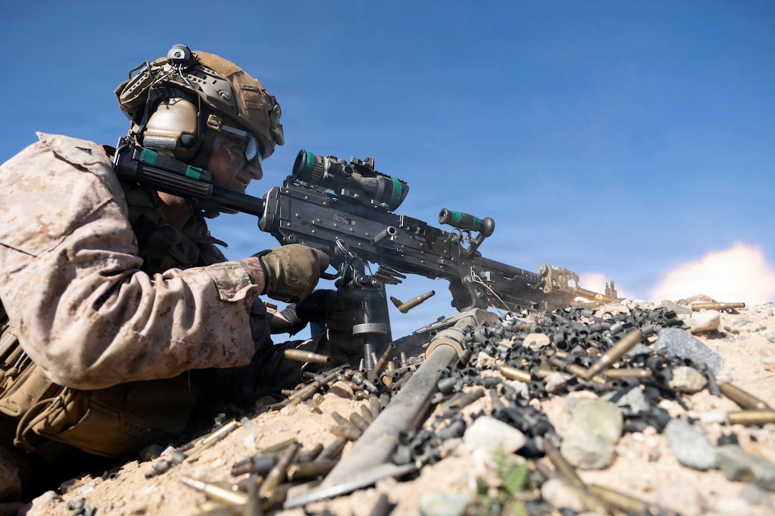 A Marine wearing a protective headset fires a machine gun while close to the ground on rocky terrain covered with shell casings.