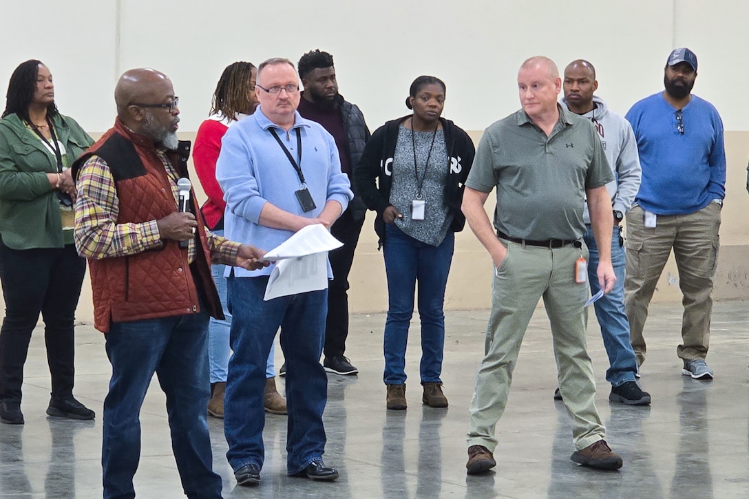 A large group of people stand around a large warehouse looking at one person who is speaking to the group.