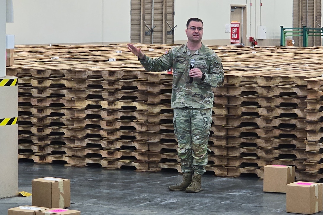 A man wearing a camouflage military uniform stands inside a large warehouse speaking into a microphone.