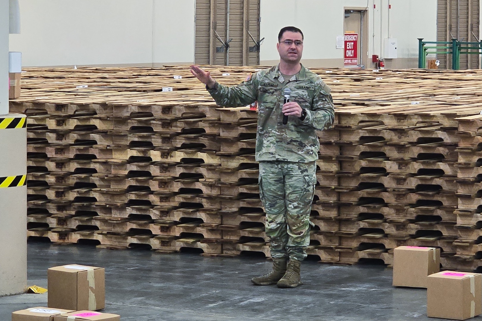 A man wearing a camouflage military uniform stands inside a large warehouse speaking into a microphone.