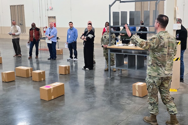 A large group of people stand around a large warehouse looking at one person who is speaking to the group.