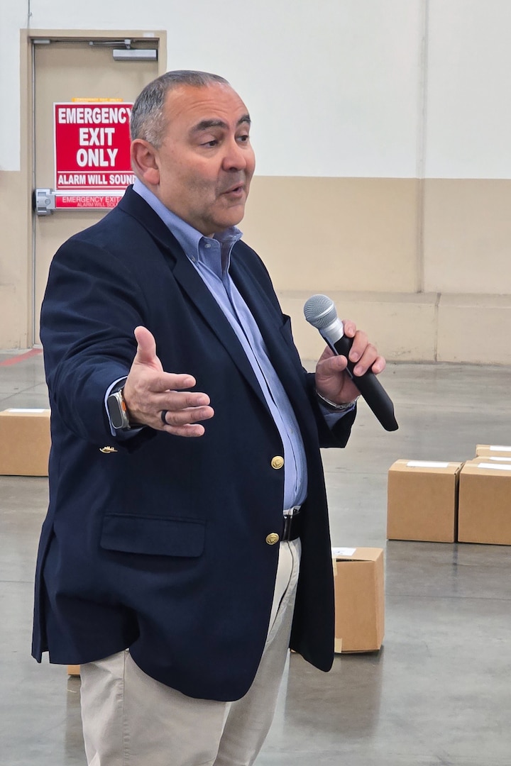 A man stands inside a large warehouse speaking into a microphone.