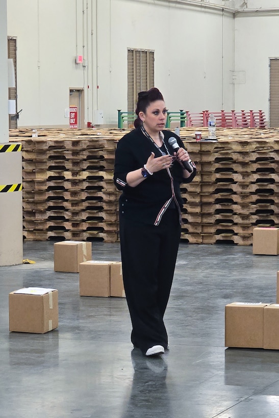 A woman stands inside a large warehouse speaking into a microphone.