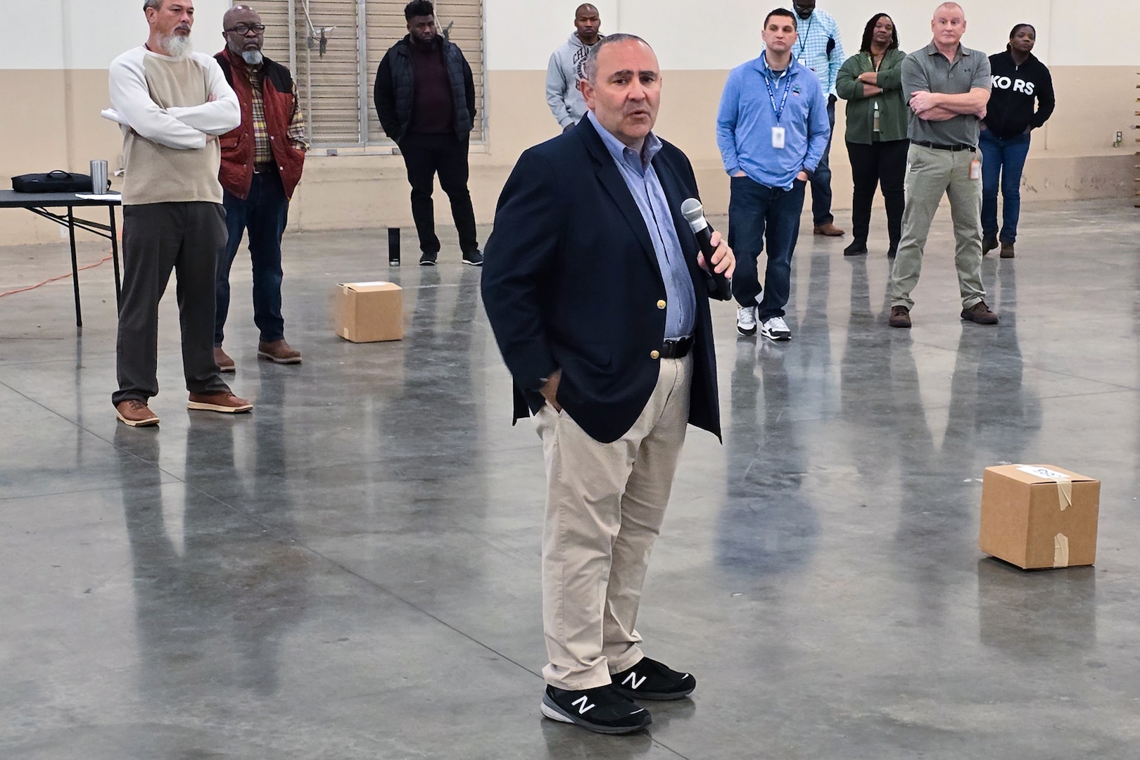 A large group of people stand around a large warehouse looking at one person who is speaking to the group.