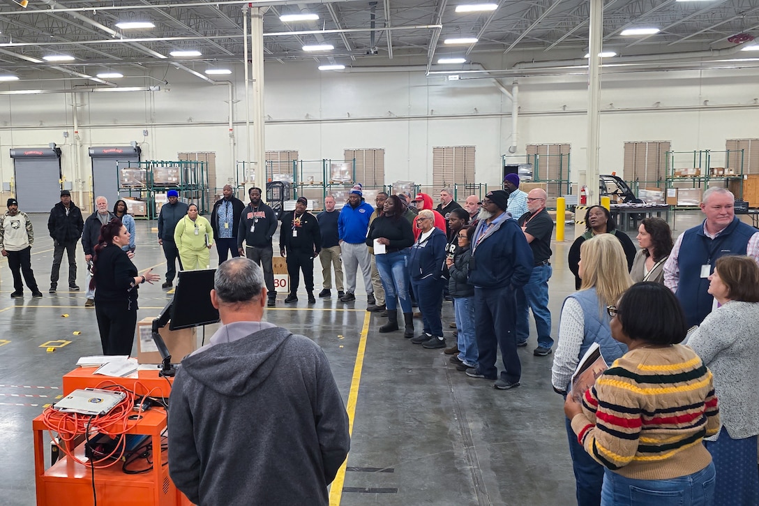 A large group of people stand around a large warehouse looking at one person who is speaking to the group.
