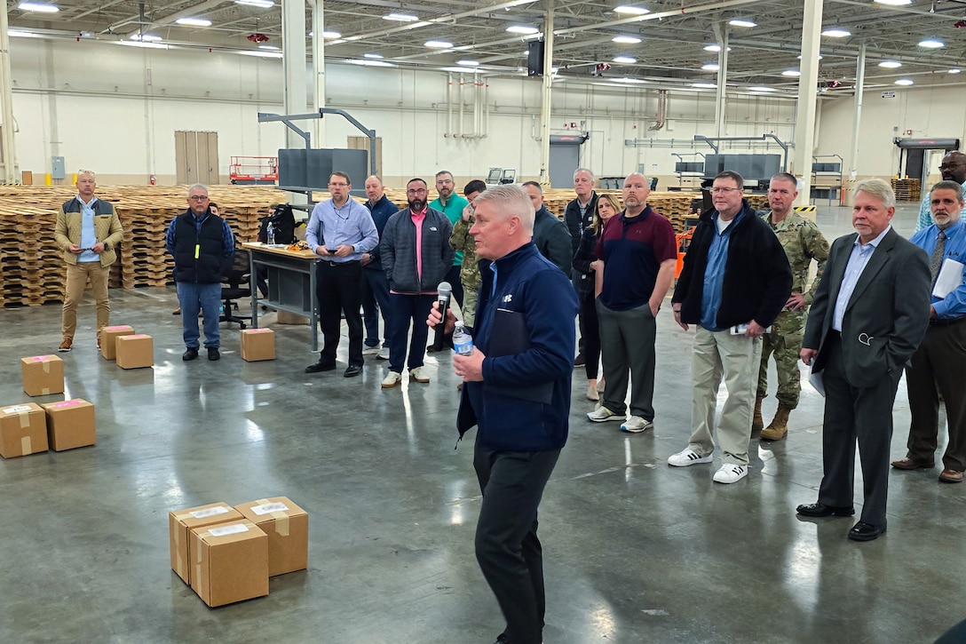 A large group of people stand around a large warehouse looking at one person who is speaking to the group.