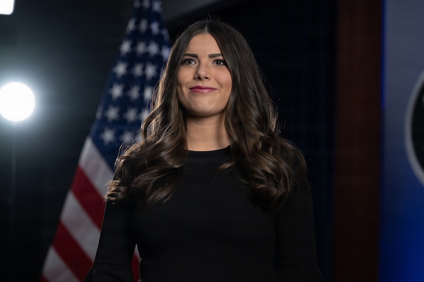 Pentagon Press Secretary Kingsley Wilson, in dark clothing, stands in front of the American flag against a dark background and studio lights.