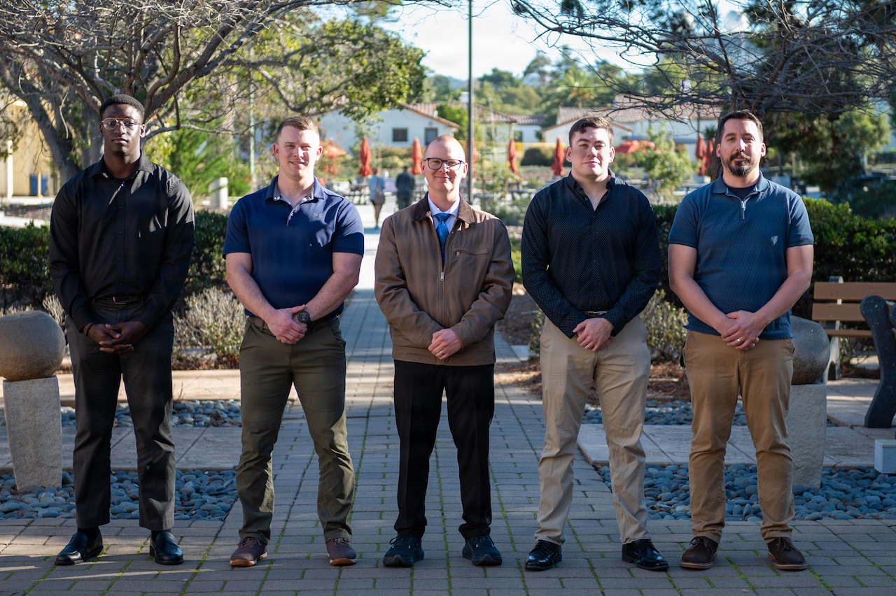 Five men dressed in casual attire stand shoulder to shoulder in a straight line while posing outdoors for a photo.
