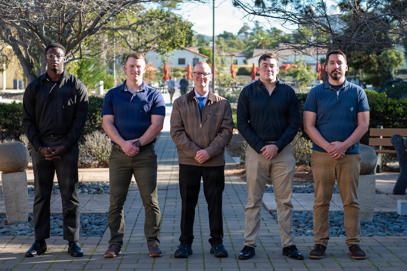 Five men dressed in casual attire stand shoulder to shoulder in a straight line while posing outdoors for a photo.