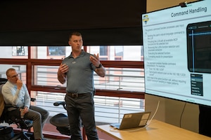 A man wearing casual business attire looks at a screen while speaking in a classroom. Another man, who is seated, watches and listens.
