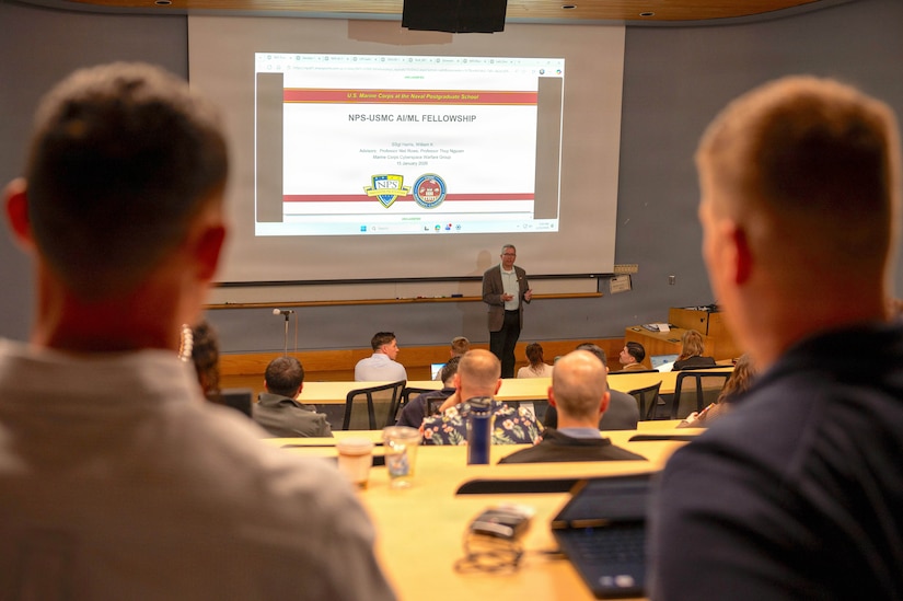 Two men with their backs to the camera stand in the back of a lecture hall watching a man with a projection screen behind him speak. There are also people seated at desks in the hall.