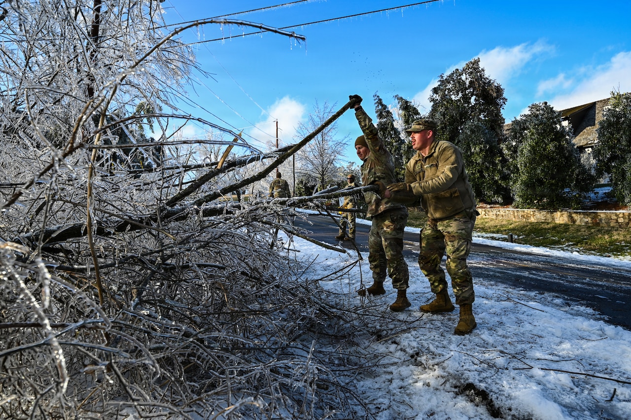 Two men in camouflage military uniforms stand outside on the side of an icy road, while working to push frozen tree branches off to the side.