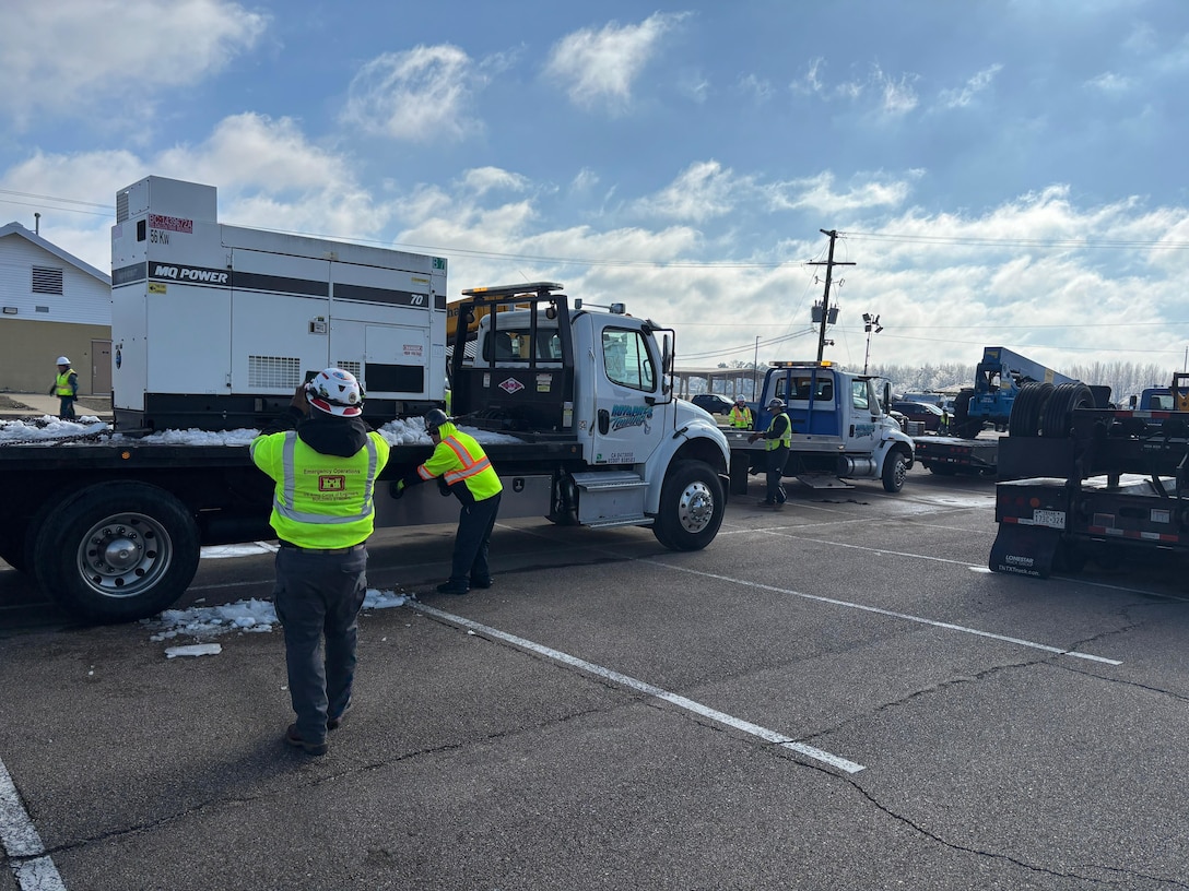 a generator is on a flatbed truck, on man is taking pictures of it while another works on the tiedown straps.
