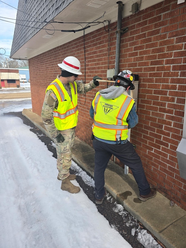 a soldier and a man look at an outdoor electrical panel of a building.