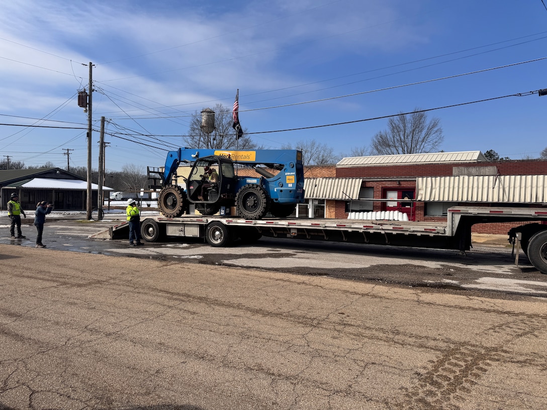 A blue forklift sits on the back of a flatbed trailer. A man sits in the cab of the forklift as he gets ready to roll it off the trailer.