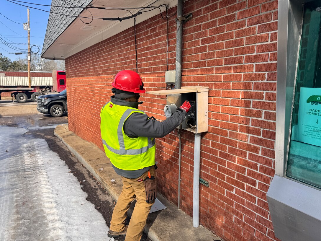 A man in a hard hat and safety vest looks at an electrical panel on the side of a red brick building.