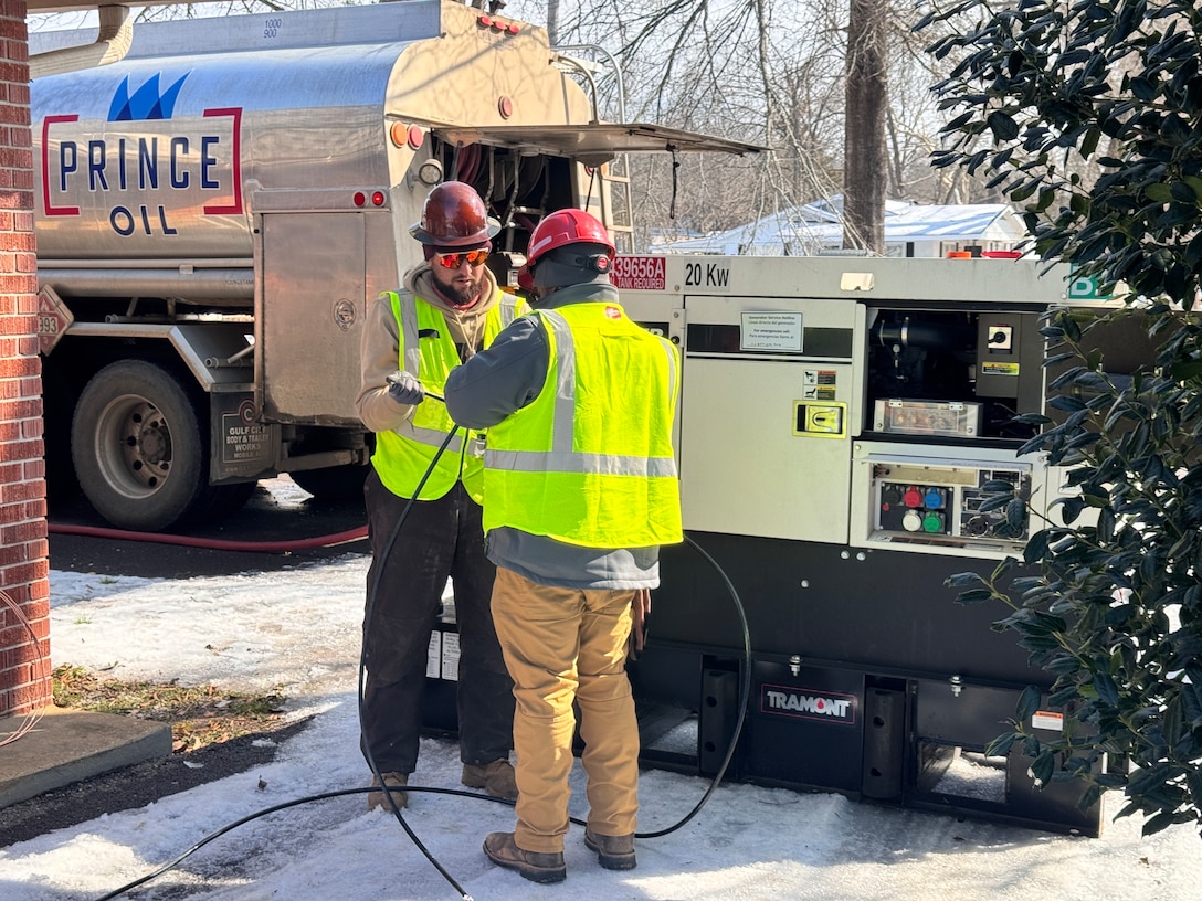 Two men work to unspool a large electrical cable. They are standing next to a large generator.