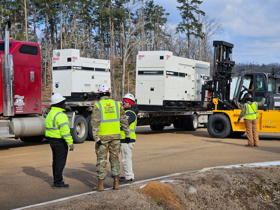 a Soldier stands with two other people as they observe a forklift remove a large generator from a flatbed truck.