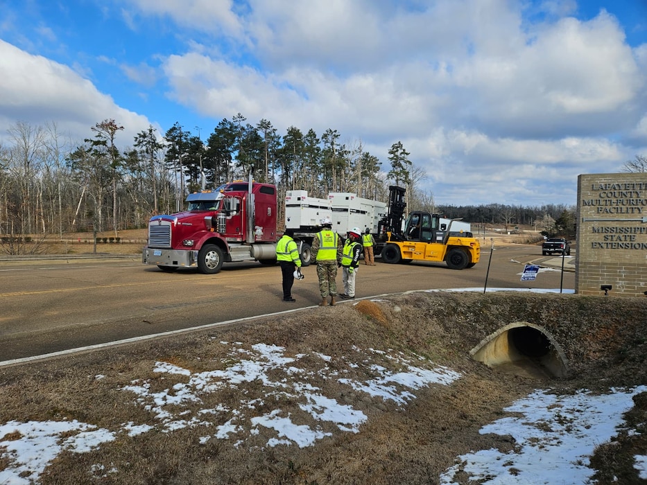 A soldier stands with two other individuals. They are all watching a forklift as it removes one of three generators from a flatbed truck.