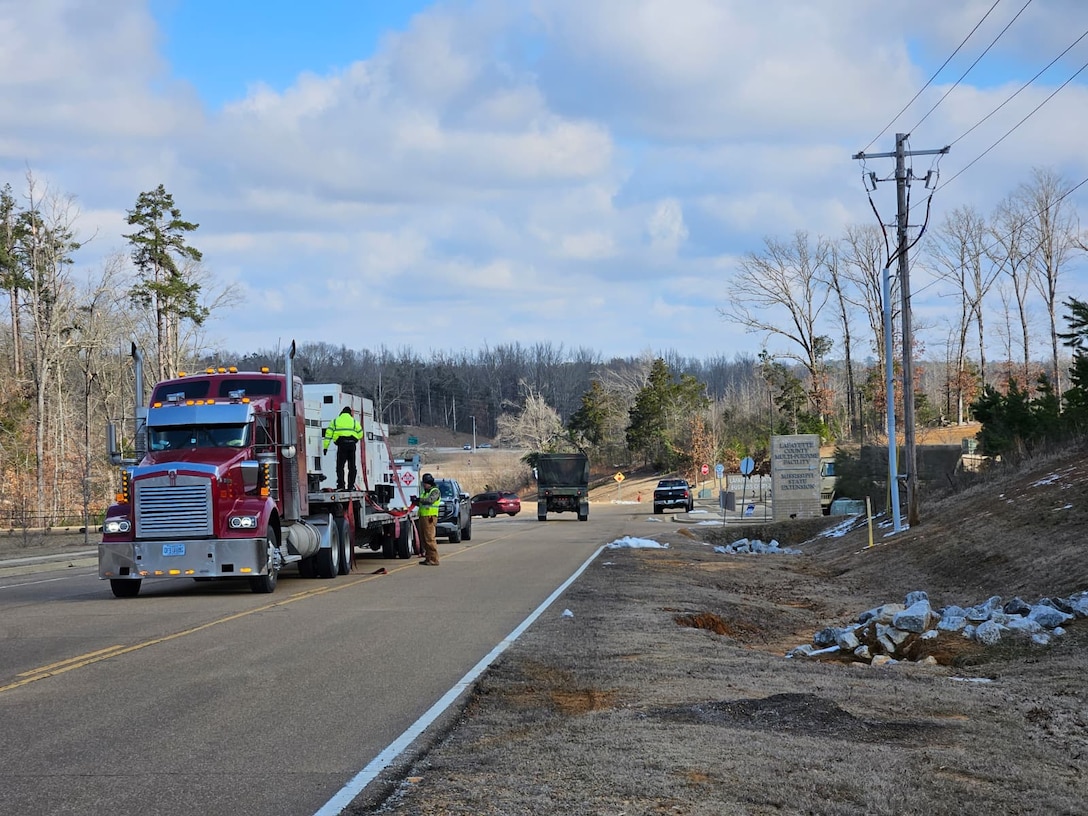 An heavy haul truck tows a large flatbed trailer with three large generators secured to it. People are working to get the generators ready for placement.