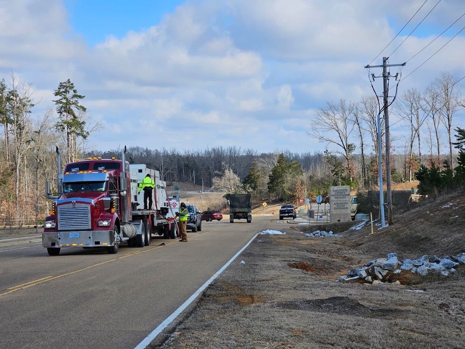 An heavy haul truck tows a large flatbed trailer with three large generators secured to it. People are working to get the generators ready for placement.