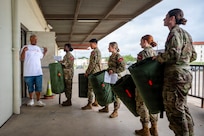 Five army soldiers stand in a line holding their newly-issued gear. They are standing outside of the warehouse. There is also a civilian employee talking to them.