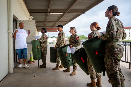 Five army soldiers stand in a line holding their newly-issued gear. They are standing outside of the warehouse. There is also a civilian employee talking to them.