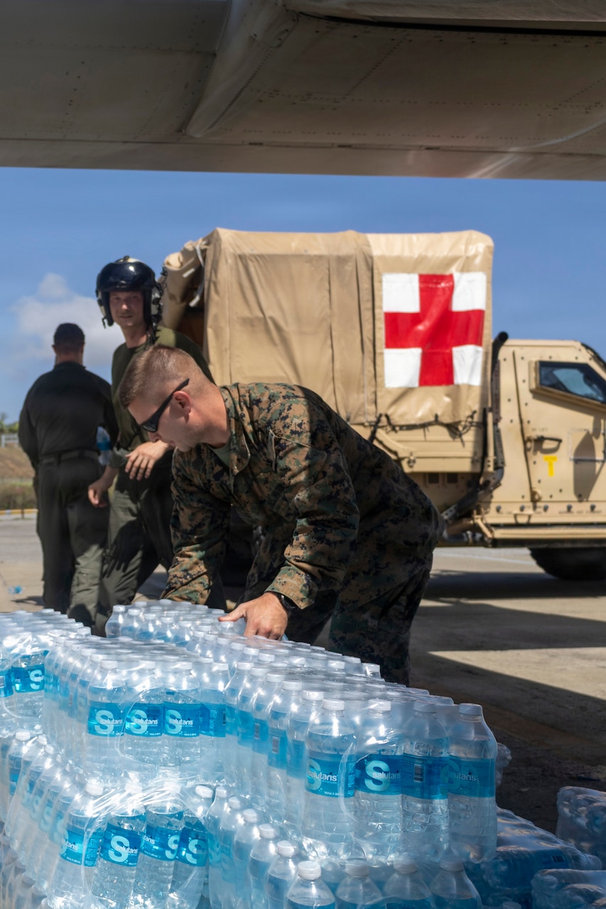 Three Marines work on a flight line, one handling cases of water and two others near a military ambulance.
