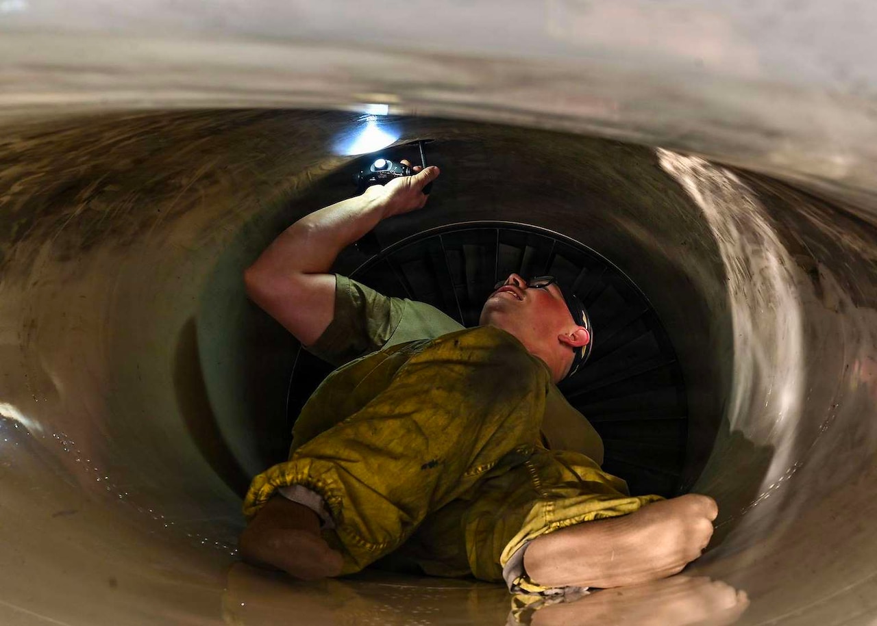 A man wearing green overalls holds a flashlight while lying on his back in an aircraft intake.