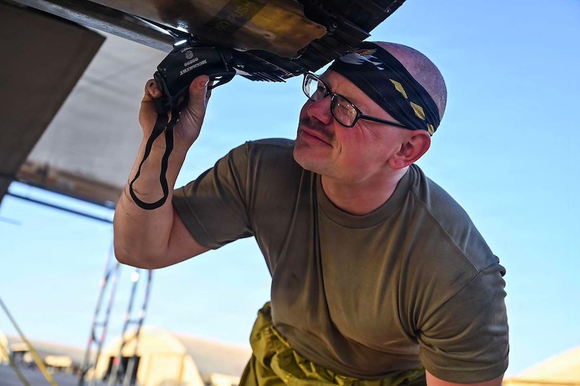 A man wearing a green shirt looks up at the underside of a military aircraft with a flashlight in his right hand.