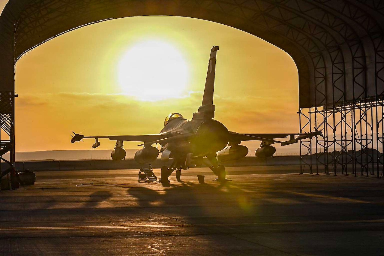 A military jet sits under a hangar on a flight line as the sun sets in front of it.