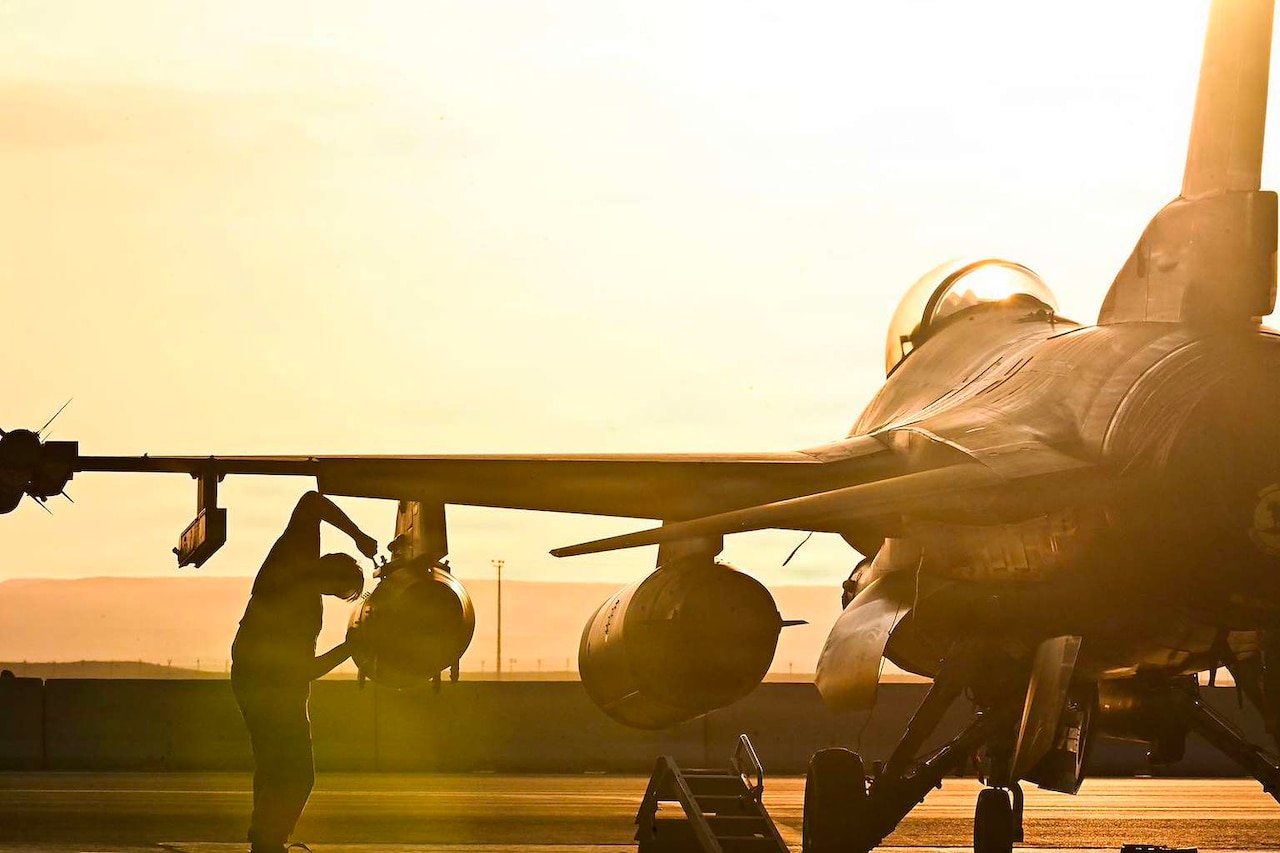 The silhouette of a man inspecting a military aircraft as the sun sets in the background.