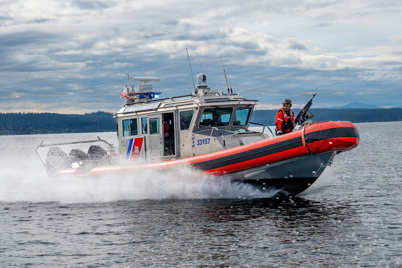 A coast guardsman wearing an orange jacket and helmet mans a machine gun on a gray boat with orange gunwhales cruises in the water on a cloudy day.