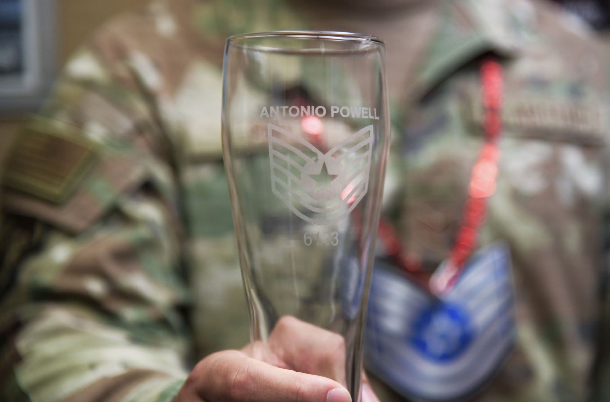 A glass etched with a name and a Staff Sergeant emblem is held by a hand.