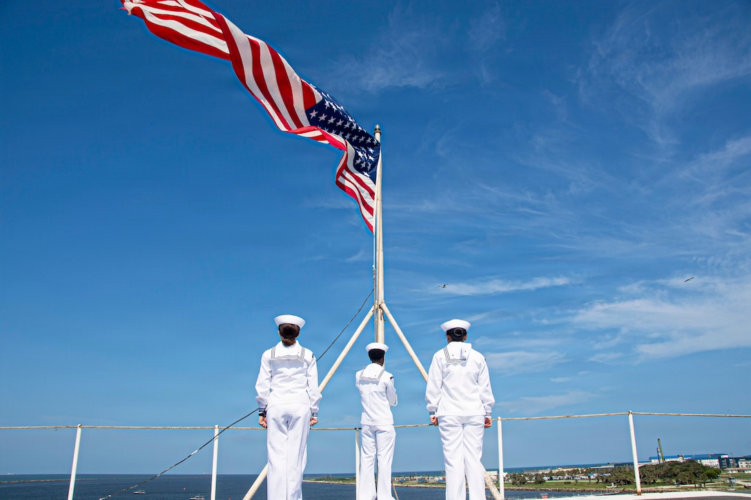 Three sailors in white uniforms stand in a triangular formation while facing an American flag as it blows in the wind aboard a ship during the day.
