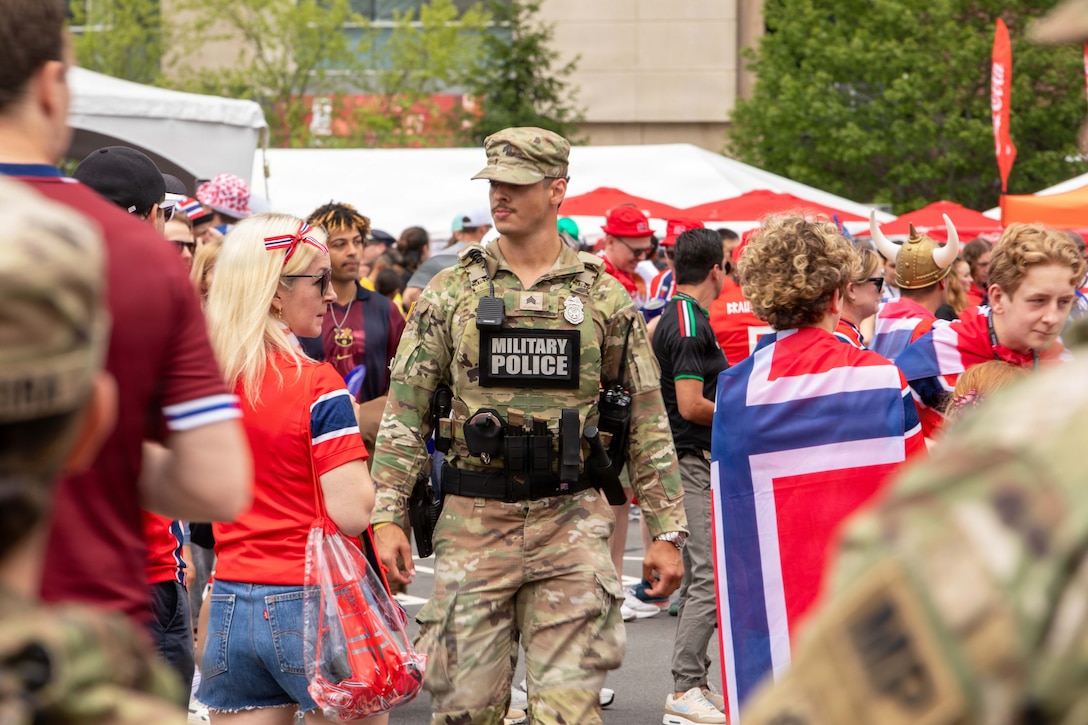 A soldier wearing police gear walks through a large crowd of soccer fans wearing red, white and blue outside of a stadium.