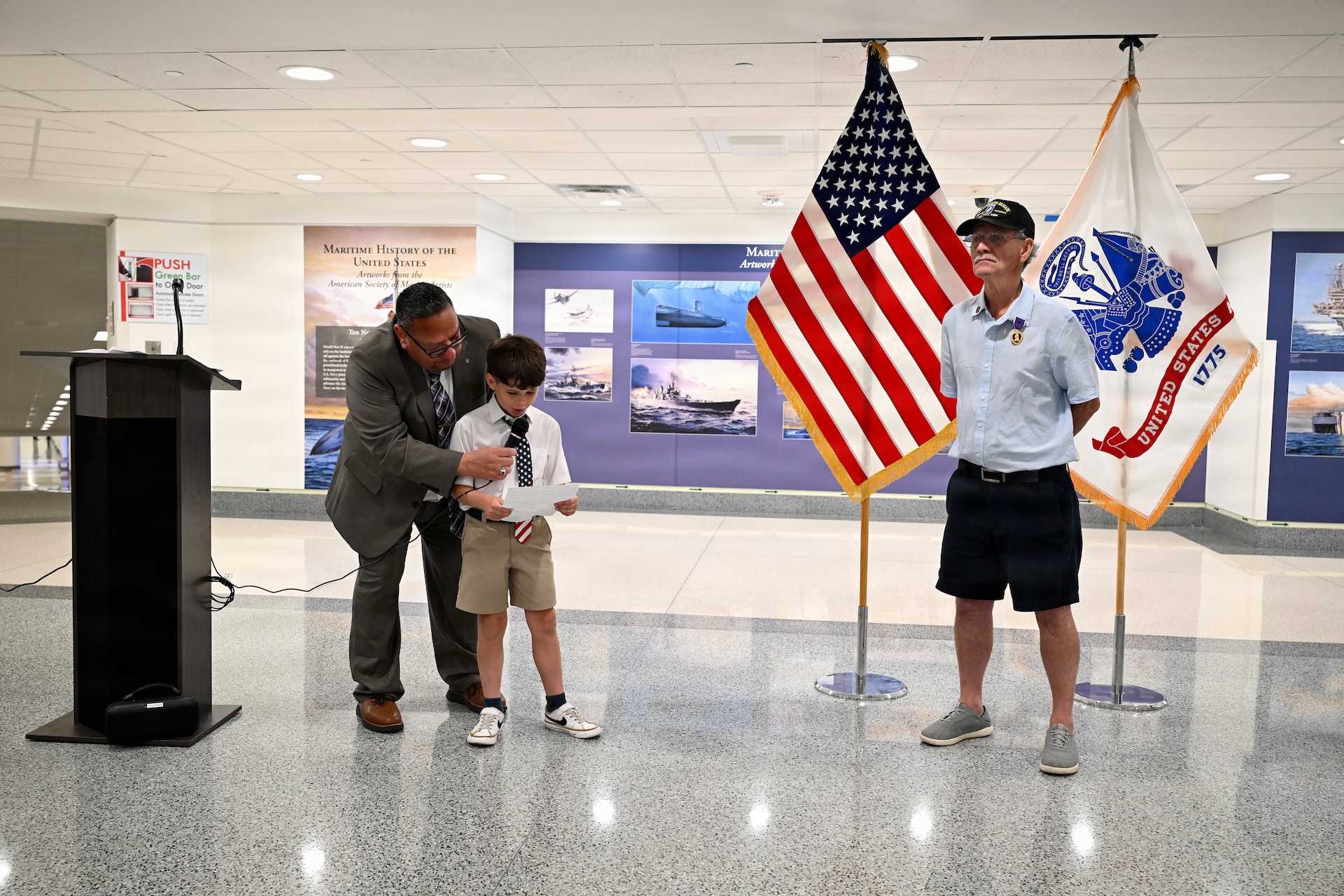 Specialist Fourth Class Raymond Steven Williams watches from the side as his eight-year-old grandson, Fynn Williams, speaks at a podium during a Purple Heart presentation ceremony at the Pentagon. Juan Rodriguez assists Fynn at the microphone.