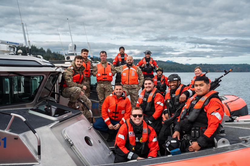 Thirteen men wearing military uniforms and life jackets pose for a photo while standing on two boats floating in the water; there are mountains in the background.