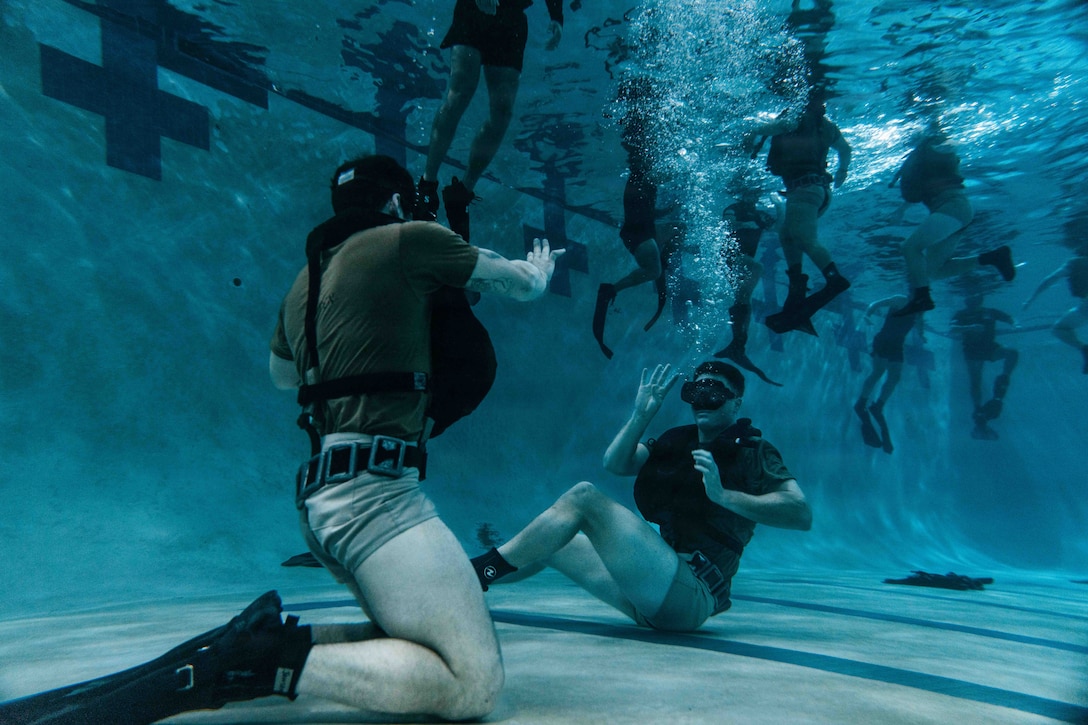 Soldiers wearing diving gear signal to each other while sitting at the bottom of a pool as nearly a dozen soldiers tread water above.