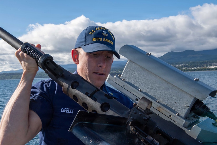 A man wearing a military uniform checks a large machine gun while on a boat in the water.