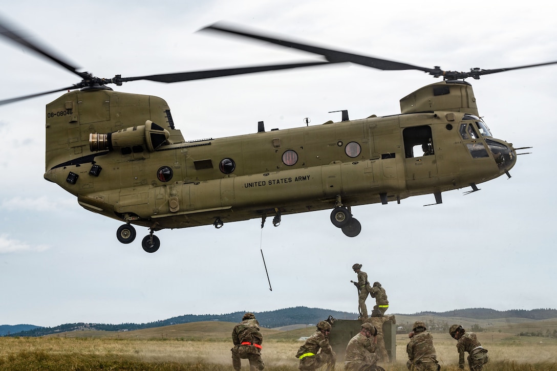 Airmen kneel next to a generator in a field as an airman standing on it prepares to attach it to hovering helicopter against a gray sky.