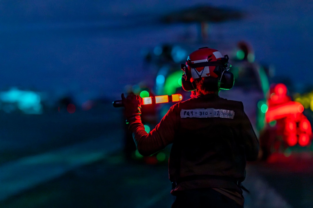 A sailor holding red batons stands in front of a helicopter in the dark illuminated by red and green lights.